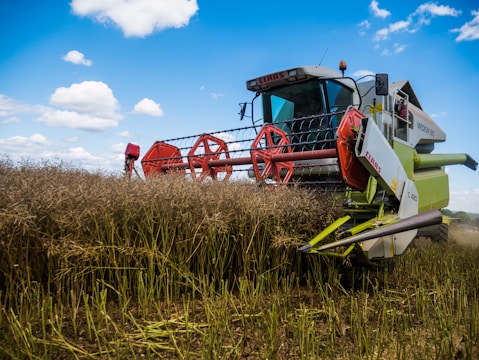 A large combine harvester is actively harvesting a field of tall crops under a partly cloudy blue sky. The machine is equipped with red rotating mechanisms and has branding on its side panel. The foreground features cut stalks, while the background shows standing crops.