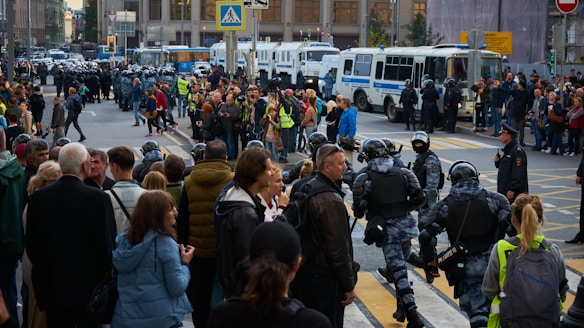 A busy urban street scene with a large group of people gathered, possibly at an event or protest. Law enforcement officers in riot gear are present, forming a line and interacting with the crowd. Several police vehicles are parked nearby, and the atmosphere appears tense and lively.