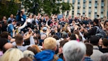 A large crowd of people gathers around, many holding cameras and smartphones, capturing the moment. In the center, several individuals appear to be speaking or addressing the crowd, surrounded by media personnel with professional cameras. Tall buildings and greenery are visible in the background.