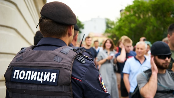 A police officer wearing a uniform with Cyrillic text stands in the foreground, with a crowd of people in the background. The crowd appears diverse in terms of age and gender, and they are gathered near a building and some greenery.