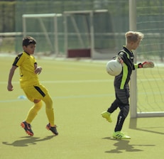 Two young boys are playing soccer on a field. One boy, dressed in yellow, is positioned to the left and seems to be getting ready to move towards the ball. The other boy, dressed as a goalkeeper in black with neon green stripes, is holding a soccer ball and is near a goalpost. The field is grassy and there are other soccer nets visible in the background.