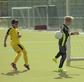 Two young boys are playing soccer on a field. One boy, dressed in yellow, is positioned to the left and seems to be getting ready to move towards the ball. The other boy, dressed as a goalkeeper in black with neon green stripes, is holding a soccer ball and is near a goalpost. The field is grassy and there are other soccer nets visible in the background.