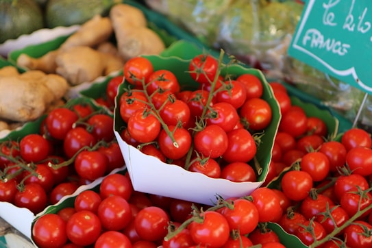 A vibrant display of fresh red tomatoes on the vine, presented in white cardboard containers. They are surrounded by other vegetables, including ginger roots, with a few greenish squash visible in the background. A green sign with white writing suggests a market or grocery setting.