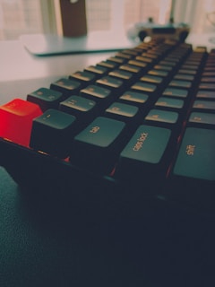 A close-up angle of a mechanical keyboard with focus on the dark black keys and a glowing red keycap on the escape key. The soft lighting creates a cozy, slightly dim ambiance. The surface below the keyboard has a smooth texture, and a blurred background includes a window and a faint silhouette of additional objects.