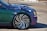 A close-up view of a car's front side featuring custom wheels and a metallic paint job. The wheel design is highly intricate, resembling a swirling pattern with chrome and dark accents. The car's body paint includes stylized flame decals in blue and white tones.
