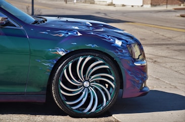 A close-up view of a car's front side featuring custom wheels and a metallic paint job. The wheel design is highly intricate, resembling a swirling pattern with chrome and dark accents. The car's body paint includes stylized flame decals in blue and white tones.