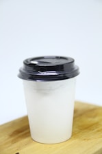 A disposable coffee cup with a black plastic lid rests on a wooden surface against a plain light background. The cup is white and appears to be made from paper.