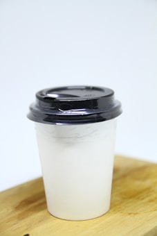 A disposable coffee cup with a black plastic lid rests on a wooden surface against a plain light background. The cup is white and appears to be made from paper.