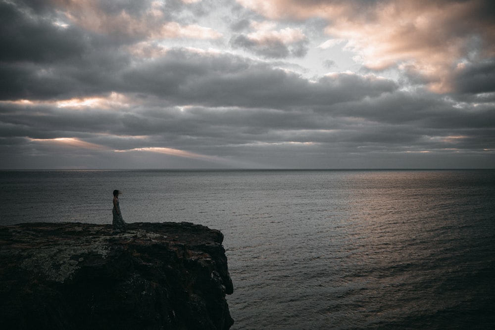 silhouette di persona in piedi sulla formazione rocciosa vicino al mare durante il giorno