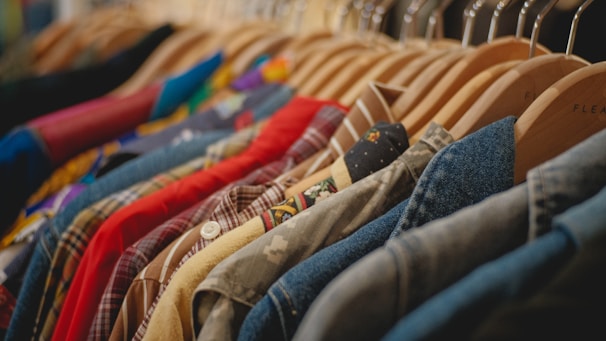 A row of various colorful clothes on wooden hangers, including plaid shirts, denim jackets, and a variety of patterns and textures. The garments are closely packed and hung on a metal rail.