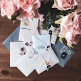 An arrangement of beautifully decorated envelopes lies on a wooden table. Each envelope features stamps and delicate floral designs or embellishments. In the upper left corner, a bouquet of pink roses adds a touch of elegance to the scene.