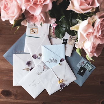 An arrangement of beautifully decorated envelopes lies on a wooden table. Each envelope features stamps and delicate floral designs or embellishments. In the upper left corner, a bouquet of pink roses adds a touch of elegance to the scene.