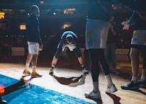 Basketball players are on a court, engaging in what appears to be a warm-up or team introduction session. The lighting is dramatic, casting strong shadows on the wooden floor. A player in the middle is stretching while others stand around him in sports attire.