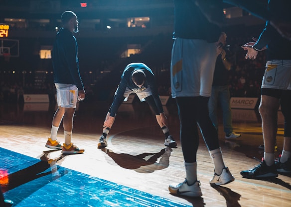 Basketball players are on a court, engaging in what appears to be a warm-up or team introduction session. The lighting is dramatic, casting strong shadows on the wooden floor. A player in the middle is stretching while others stand around him in sports attire.