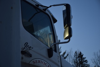 A close-up of a white truck featuring large side mirrors reflecting the evening light, with trees visible in the background. The name 'Joe' is written on the side of the door, and the word 'SERVICE' is partially visible below it.