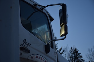 A close-up of a white truck featuring large side mirrors reflecting the evening light, with trees visible in the background. The name 'Joe' is written on the side of the door, and the word 'SERVICE' is partially visible below it.