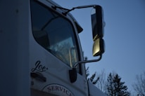 A close-up of a white truck featuring large side mirrors reflecting the evening light, with trees visible in the background. The name 'Joe' is written on the side of the door, and the word 'SERVICE' is partially visible below it.