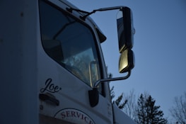 A close-up of a white truck featuring large side mirrors reflecting the evening light, with trees visible in the background. The name 'Joe' is written on the side of the door, and the word 'SERVICE' is partially visible below it.