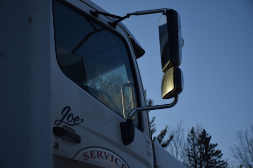 A close-up of a white truck featuring large side mirrors reflecting the evening light, with trees visible in the background. The name 'Joe' is written on the side of the door, and the word 'SERVICE' is partially visible below it.
