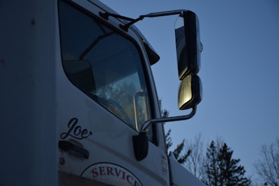 A close-up of a white truck featuring large side mirrors reflecting the evening light, with trees visible in the background. The name 'Joe' is written on the side of the door, and the word 'SERVICE' is partially visible below it.