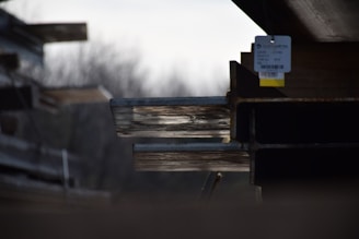 Several stacked steel beams with visible rust and a tag hanging from one. The background is blurred, creating depth and focus on the metal beams.