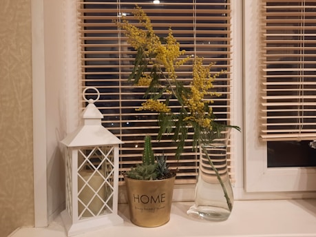 A windowsill adorned with a decorative white lantern housing a candle, a potted small green plant in a gold pot labeled 'HOME', and a clear glass vase holding sprigs of yellow flowers. The background features blinds covering a window.