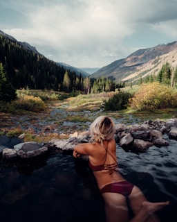 A woman with blonde hair is relaxing in a natural hot spring pool, surrounded by a serene landscape of mountain ranges and dense forests. The foreground features rocks lining the edge of the pool, while the background reveals lush greenery and towering peaks beneath a cloudy sky.