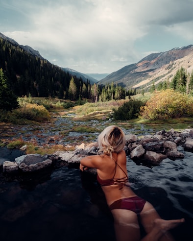A woman with blonde hair is relaxing in a natural hot spring pool, surrounded by a serene landscape of mountain ranges and dense forests. The foreground features rocks lining the edge of the pool, while the background reveals lush greenery and towering peaks beneath a cloudy sky.