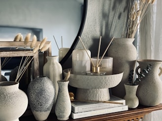 Aesthetic arrangement of various ceramic vases and decor pieces on a wooden surface. The setup includes different textured and shaped vases, some of which hold dried grass stems. There are also candles and hardcover books adding to the decoration. The background features a large round mirror, enhancing the stylish and modern vibe.