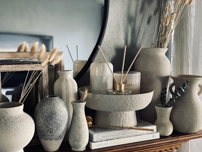 Aesthetic arrangement of various ceramic vases and decor pieces on a wooden surface. The setup includes different textured and shaped vases, some of which hold dried grass stems. There are also candles and hardcover books adding to the decoration. The background features a large round mirror, enhancing the stylish and modern vibe.