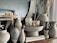 Aesthetic arrangement of various ceramic vases and decor pieces on a wooden surface. The setup includes different textured and shaped vases, some of which hold dried grass stems. There are also candles and hardcover books adding to the decoration. The background features a large round mirror, enhancing the stylish and modern vibe.