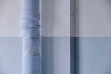 A vertical pipe wrapped in layers of light-colored material runs alongside a two-toned wall with visible cracks and a subtle texture. The wall features a horizontal division between off-white and light blue sections, creating an industrial or minimalist aesthetic.