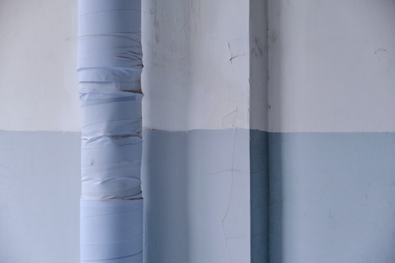 A vertical pipe wrapped in layers of light-colored material runs alongside a two-toned wall with visible cracks and a subtle texture. The wall features a horizontal division between off-white and light blue sections, creating an industrial or minimalist aesthetic.