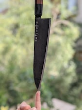 A hand is balancing a large kitchen knife on the tip of a finger against a blurred green background, demonstrating precision and skill. The knife has a black blade and a wooden handle, with some characters engraved on the blade.