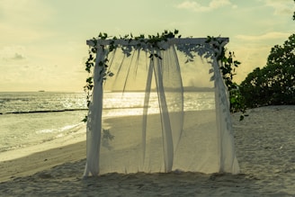 A beach setting featuring a sheer, white canopy decorated with green leaves, positioned in the sand. The ocean is in the background, reflecting the golden hues of the sunset, creating a tranquil and serene atmosphere. The sky is lightly clouded, contributing to the peaceful ambiance.