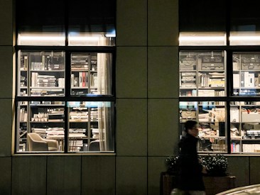 A nighttime scene featuring a well-lit window revealing a cluttered office space filled with numerous stacks of books, files, and binders. The shelves are densely packed, and a chair is visible inside. A blurred figure walks past the window, moving along the sidewalk outside.
