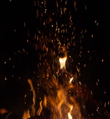 Bright, glowing flames and sparks rise against a dark background, creating a dynamic and intense visual. The flames vary in intensity, with some forming distinct shapes while others blur into the darkness. The sparks add texture and movement to the scene.