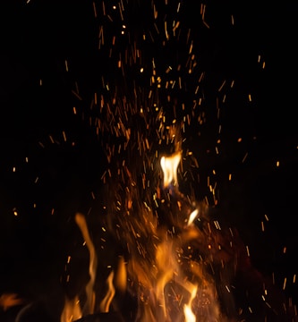 Bright, glowing flames and sparks rise against a dark background, creating a dynamic and intense visual. The flames vary in intensity, with some forming distinct shapes while others blur into the darkness. The sparks add texture and movement to the scene.