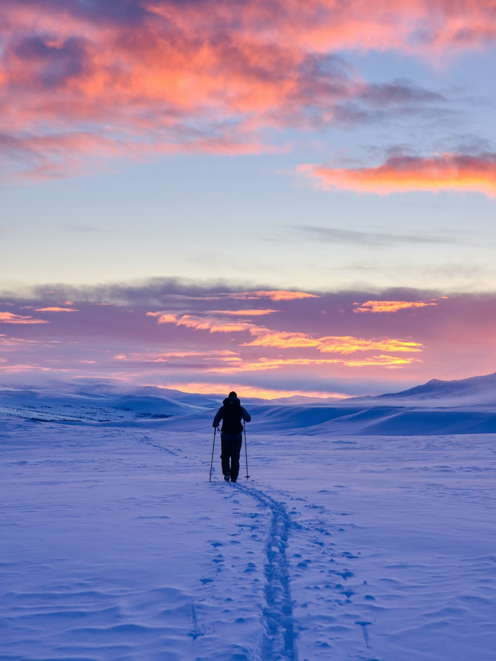 homem em jaqueta preta andando no chão coberto de neve durante o pôr do sol