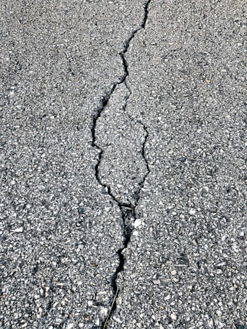 A close-up view of a cracked asphalt surface, showing a prominent crack running vertically through the rough, textured pavement.