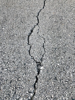 A close-up view of a cracked asphalt surface, showing a prominent crack running vertically through the rough, textured pavement.
