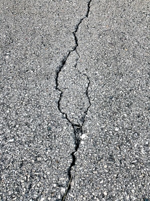A close-up view of a cracked asphalt surface, showing a prominent crack running vertically through the rough, textured pavement.