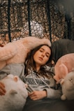 A person is lounging comfortably on a plush chair surrounded by large, soft stuffed animals. They are wearing a gray hoodie with 'Brooklyn' written on it. The background includes a woven pattern that adds texture to the scene.