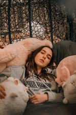 A person is lounging comfortably on a plush chair surrounded by large, soft stuffed animals. They are wearing a gray hoodie with 'Brooklyn' written on it. The background includes a woven pattern that adds texture to the scene.