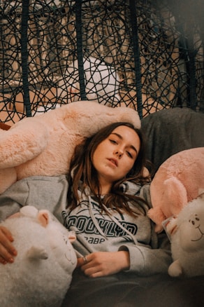A person is lounging comfortably on a plush chair surrounded by large, soft stuffed animals. They are wearing a gray hoodie with 'Brooklyn' written on it. The background includes a woven pattern that adds texture to the scene.