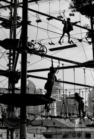 Silhouetted figures are navigating a complex ropes course high above the ground, set against an urban backdrop with buildings and trees faintly visible. The scene is dynamic and captures a sense of movement and challenge as the participants engage with various obstacles.