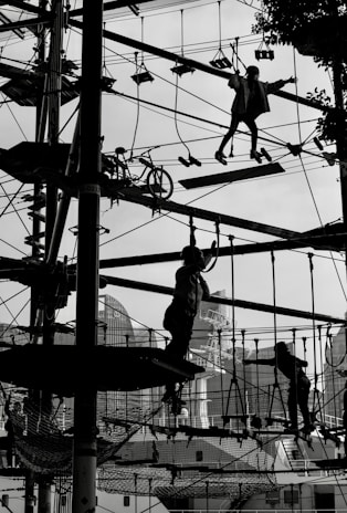 Silhouetted figures are navigating a complex ropes course high above the ground, set against an urban backdrop with buildings and trees faintly visible. The scene is dynamic and captures a sense of movement and challenge as the participants engage with various obstacles.