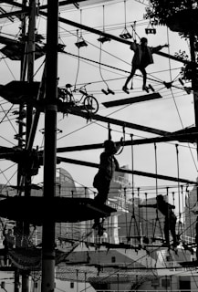 Silhouetted figures are navigating a complex ropes course high above the ground, set against an urban backdrop with buildings and trees faintly visible. The scene is dynamic and captures a sense of movement and challenge as the participants engage with various obstacles.