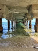 Underneath a concrete pier, pilings stretch into the distance over clear turquoise water. Sunlight filters through, casting reflections and shadows on the water and sandy shore.