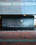 A modern building facade with a large stone wall displaying the name 'National Museum of American Jewish History, Lyn and George Ross Building.' The ground is covered with red and gray brick tiles, and the upper section features reflective glass panels.